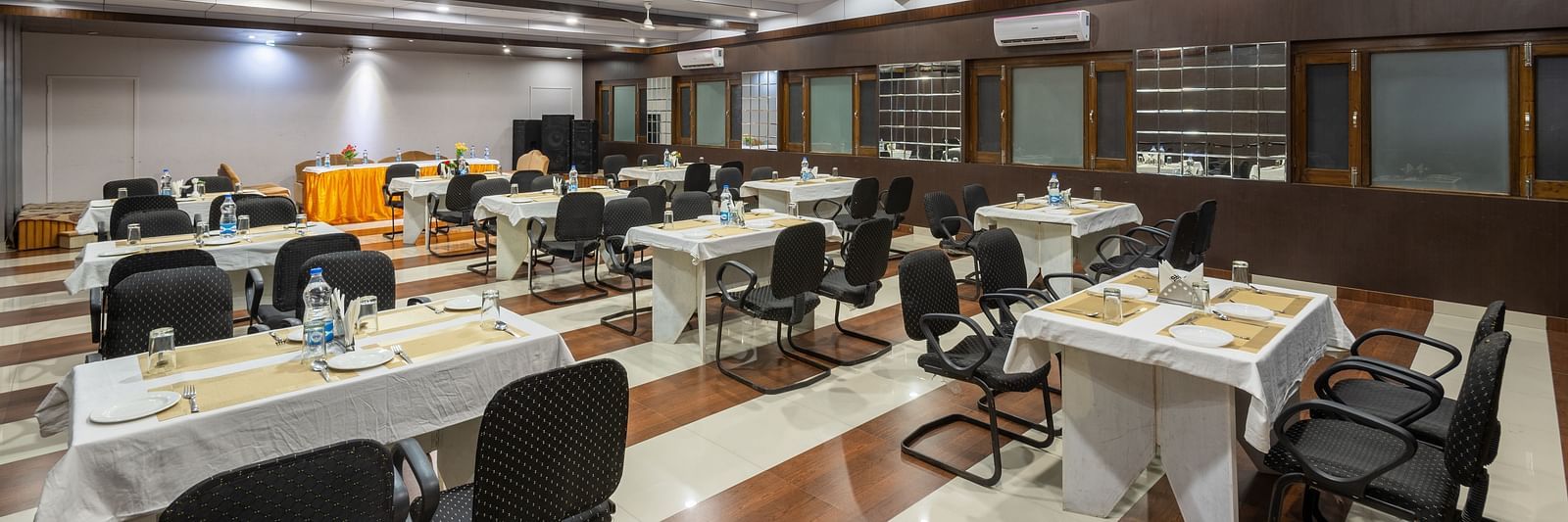 A well-lit conference room with rows of tables and chairs, featuring a projector screen and a fan - DLS Meghavan Resort